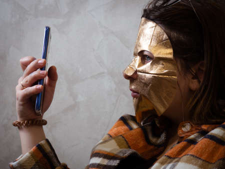 brunette girl with a mobile phone in a gold sheet mask on her face. relax in an armchair during beauty treatments at home on an armchair.の写真素材