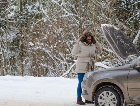 beautiful girl near the engine of a broken car on a winter snowy road. freeze and calls for help