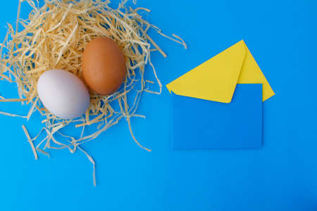 A brown and white chicken egg and a small red one on hay on a blue background. Next to it is a yellow and blue envelope. Easter card conceptの写真素材