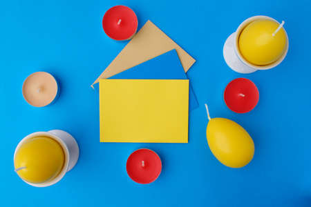 Two yellow candles in the form of an egg in a stand on a blue background. In the center are colored envelopes. Nearby are small tea candles. Easter card conceptの写真素材