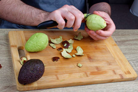 There is a knife on a wooden cutting board, next to two avocados. A man peels a third avocado.の写真素材