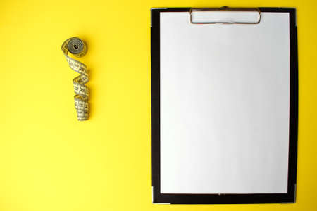Notepad with a white sheet of paper for notes, next to it twisted centimeter on a yellow background. To-do list. Copy space. Healthy food and diet concept.の写真素材