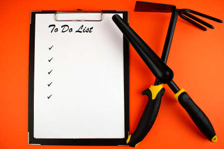 To do list. Notepad with a white sheet of paper, next to the garden shovel and hoe on an orange background. Gardening concept. Copy space. Flat lay.の写真素材