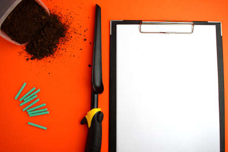Notepad with a white sheet of paper, next to the ground, garden shovel and green fertilizer on an orange background. Gardening concept. Copy space. Flat lay.の写真素材