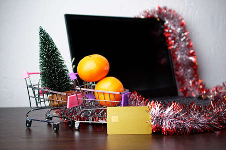Shopping baskets with a Christmas tree, tangerines and a bank card, on background tinsel and laptop.の写真素材