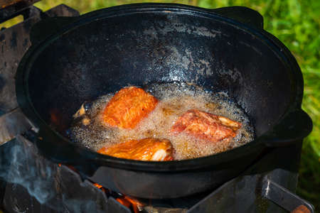 Close-up in a black cast-iron cauldron on the fire boils vegetable oil and tasty pieces of pork ribs are fried. food in nature. Small depth of field.の写真素材
