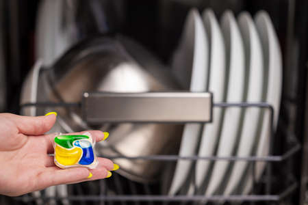 Close-up of young women hand holding a colored capsule for the dishwasher. In the background, out of focus, is a dishwasher with clean dishes. Small depth of field.の写真素材