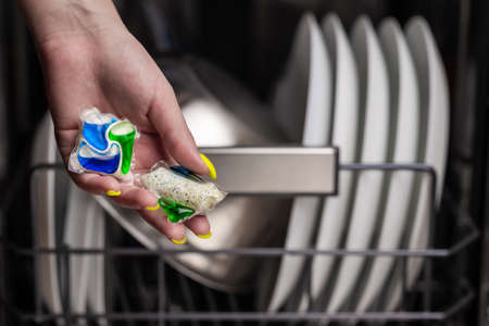Close-up of young women hand holding two colored capsule for the dishwasher. In the background, out of focus, is a dishwasher with clean dishes. Small depth of field.の写真素材