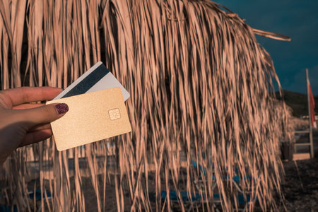 Golden and White Bank Card In Woman Hand On Background Of Beach Umbrellas Made Of Palm Leaves In Moraitika, Corfu, Greece. The Concept Of Payment For Relax And Unlimited Possibilities. Copy Space.の写真素材
