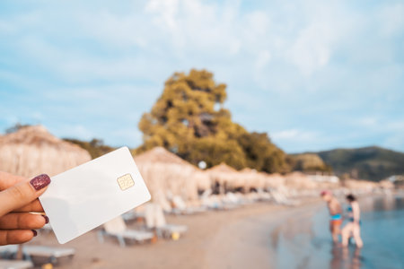 White Bank Card In Woman Hand On Background Of Hotel Beach, Family Comes Out Of Sea, Moraitika, Corfu, Greece. The Concept Of Payment For Vacation. Copy Space.の写真素材