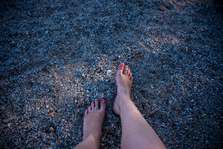 Close-Up Of Female Legs With Bright Acid Pedicure Against Background Of Pebble Beach In Moraitika, Corfu, Greece. Dark Key Shotの写真素材
