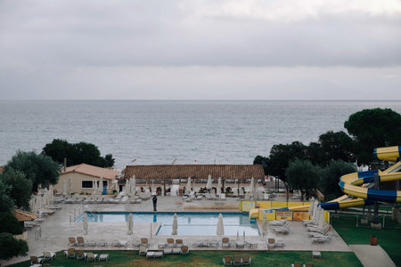 Moraitika, Greece - 09 27 2022: View on Ionian Sea and Greece Balkan Peninsula, Empty Swimming Pool, Water Slide And Sunbeds in Hotel, Due To Bad Weather And Rain. Worker Is Cleaning Pool.のeditorial素材