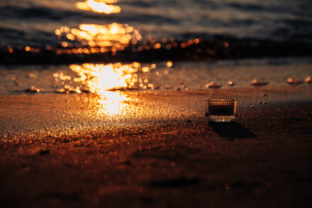 Golden Rays Of Sun Are Reflected In Light Waves, Creating Dancing Reflections On Sand And Water. Candlestick Lies On Sand. Sunrise On Beach Is Considered Romantic Time.の写真素材