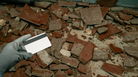womans hand adorned in construction gloves, holding a bank card, set against a backdrop of shattered tiles on the floor. Symbolizing the initiation of renovation and the considerable expenses associated with repairs.の写真素材