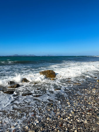 Waves Crash Against Rocks Along the Shore Under a Clear Blue Sky in a Coastal Landscape Settingの写真素材