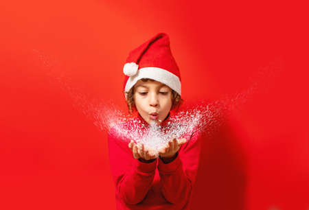 Cute kid dressing a Santa Claus hat blowing wind on his hand. Centered subject with copy space on the red backgroundの写真素材