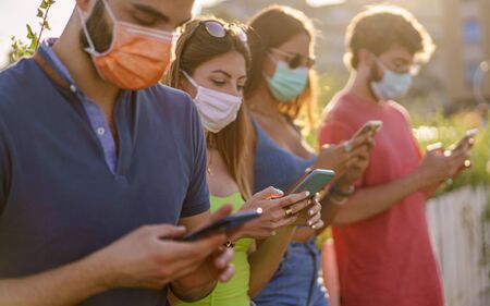 Group of friends wearing safety mask for coronavirus pandemic using smartphones. Focus on one woman's hands.の写真素材