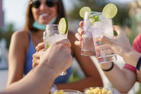 Friends drinking cocktails in an outdoors snack bar restaurant in the summer wearing face mask on to be protected from coronavirus - Happy people cheering with mojito and having funの写真素材
