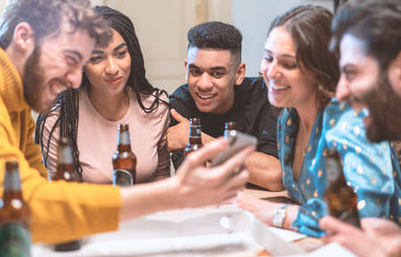 Group of smiling young people watching video on smartphone and drinking beers in a house. Multiracial friends having fun using smartphone, social network apps and new technologies concept.の写真素材