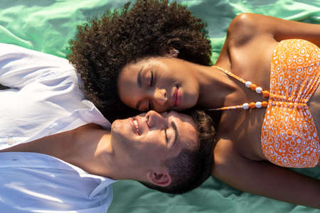 Two young people lying down cheek to cheek on a beach towel with eyes closed. Interracial concept of mixed race couple on vacation.の写真素材