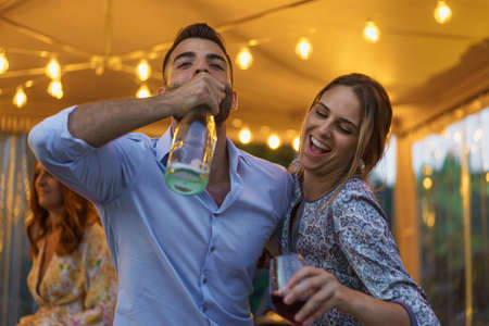 Young couple drinking sparkling white wine from the bottle during a night event party. People having fun drinking alcoholic drinks concept.の写真素材