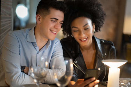 An interracial couple of young people sitting on a restaurant table and watching social network contents on the smartphone. People using tech concept.の写真素材