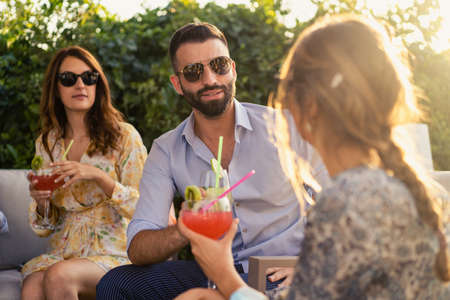 Group of young people talking together having fun while drinking soft drinks and find themselves together sitting on a sofa outdoors in the garden.の写真素材