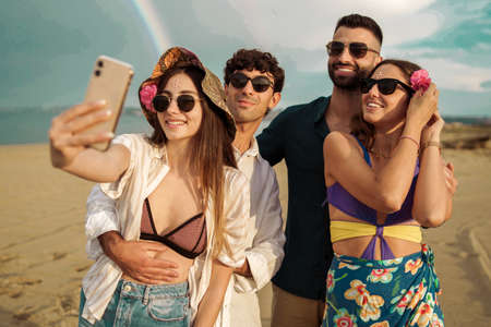 Group of friends taking a selfie on the beach against a beautiful rainbowの写真素材