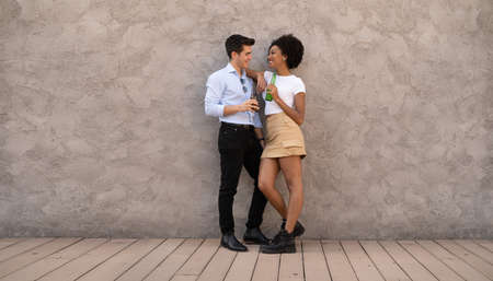 interracial couple of young people standing against a wall flirting, talking and drinking beers togetherの写真素材