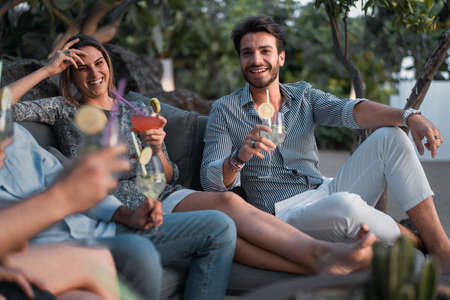Gathering of young people having fun in a cocktail party sitting on a sofa in a terrace. Friends celebrating lifestyle concept.の写真素材
