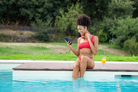 An African-American girl sitting on the edge of a swimming pool smiles while video-calling with her smartphone while holding a glass of fruit juice.の写真素材