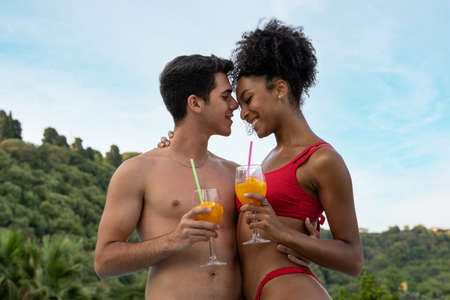 Multiracial young couple holding fruit juice glasses tenderly touching their foreheads. Surrounded by nature and trees on background.の写真素材