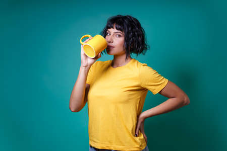 A young woman in yellow drinks from a cup of milk. Studio shot, isolated on a color background.の写真素材