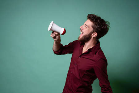 Bearded young guy talking loud in a megaphone. Concept of people raising their voices beyond the crowd.の写真素材