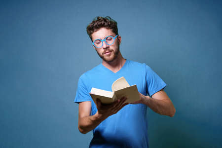 Portrait of a young student man with eyeglasses reading a book. Portrait isolated on blue colored background. Studio shot.の写真素材