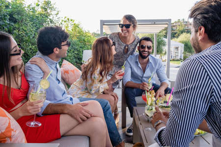 A group of laughing young people gathers at the aperitif party on the terrace. Millennials congregated in the garden joke together.の写真素材