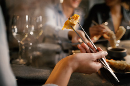Detail of a woman's hand grabbing a spring roll with metallic chopsticks. He sits at a table with friends in a fancy asiatic fusion food restaurant.の写真素材
