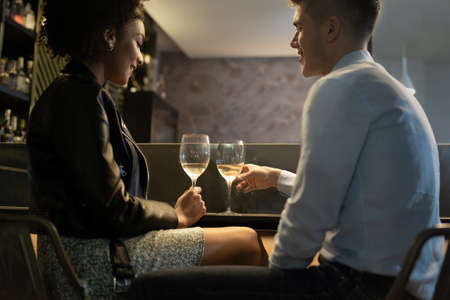 Romantic interracial couple drinking white wine sitting at the pub restaurant counter. Main focus on the wineglasses. Girlfriend and boyfriend clinking with glasses.の写真素材