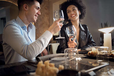 Happy Interracial couple having fun talking, eating and drinking at fusion food restaurant. Young people dating clinking wineglasses.の写真素材
