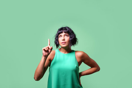 Caucasian young brunette woman pointing finger and eyes up. Isolated portrait on green background suitable for advertisement, web banners, display.の写真素材