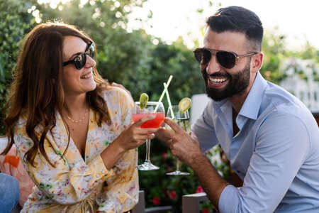 Cheerful couple of young people having fun laughing clinking tropical cocktails at a garden terrace party. Friends toasting outdoorsの写真素材