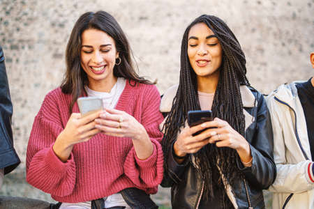 Two women best friends using smartphone together standing outdoors. Gen z young people using social network and new technology concept.の写真素材