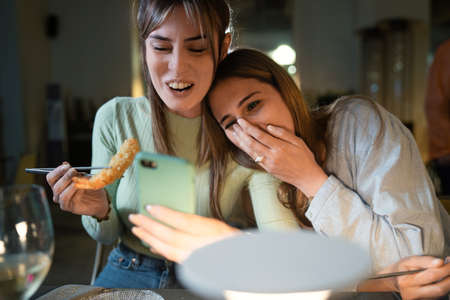 Best friends watching funny content on the smartphone screen and having fun while they're eating at Chinese restaurant. Concept of girls having fun using tech.の写真素材