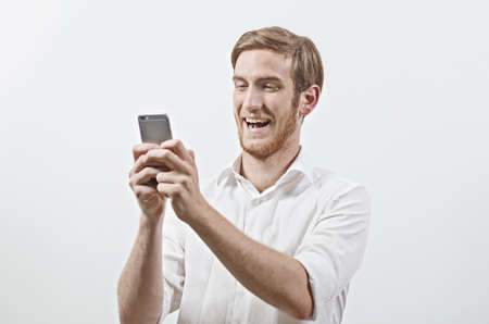 Very Happy Smiling Young Adult Man in White Shirt Looking at His Mobile Phoneの写真素材