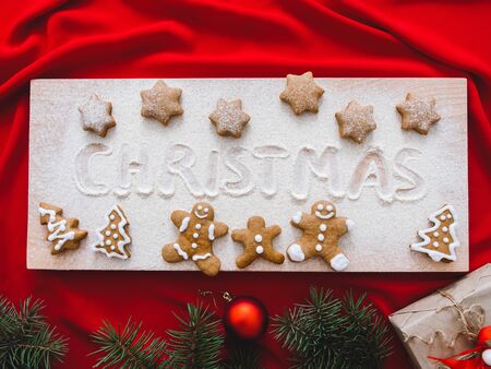 Christmas still life with traditional gingerbread cookies on wooden board. Homemade Christmas shortbread star shaped and gingerbread family sugar cookies with sugar powder, gift box, green tre, fir tree and cookie. Christmas baking on a red background. Overhead view. Veganの写真素材