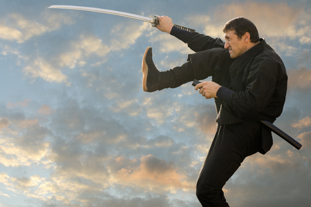 The black ninja is kicking and holding a sword on sunset cloudy sky background.の写真素材