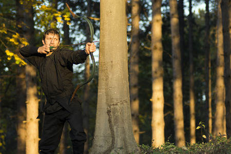 Archer in black kimono is aiming with traditional bow and arrow in the forest.