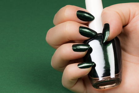Female hand with dark green nails is holding dark green nail polish on green background.の写真素材