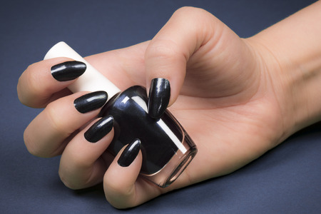 Female hand with dark nails is holding dark nail polish on blue background.の写真素材