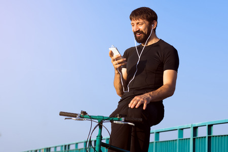The cheerful smiling cyclist with earphones is using phone outdoors.の写真素材